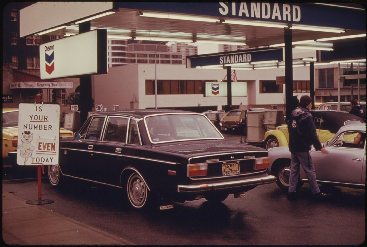 Des voitures des années 1970 attendent sous la marquise d'une station-service. Au premier plan, un écriteau sur pied porte un dessin et les mots « Is your number even today? ».
