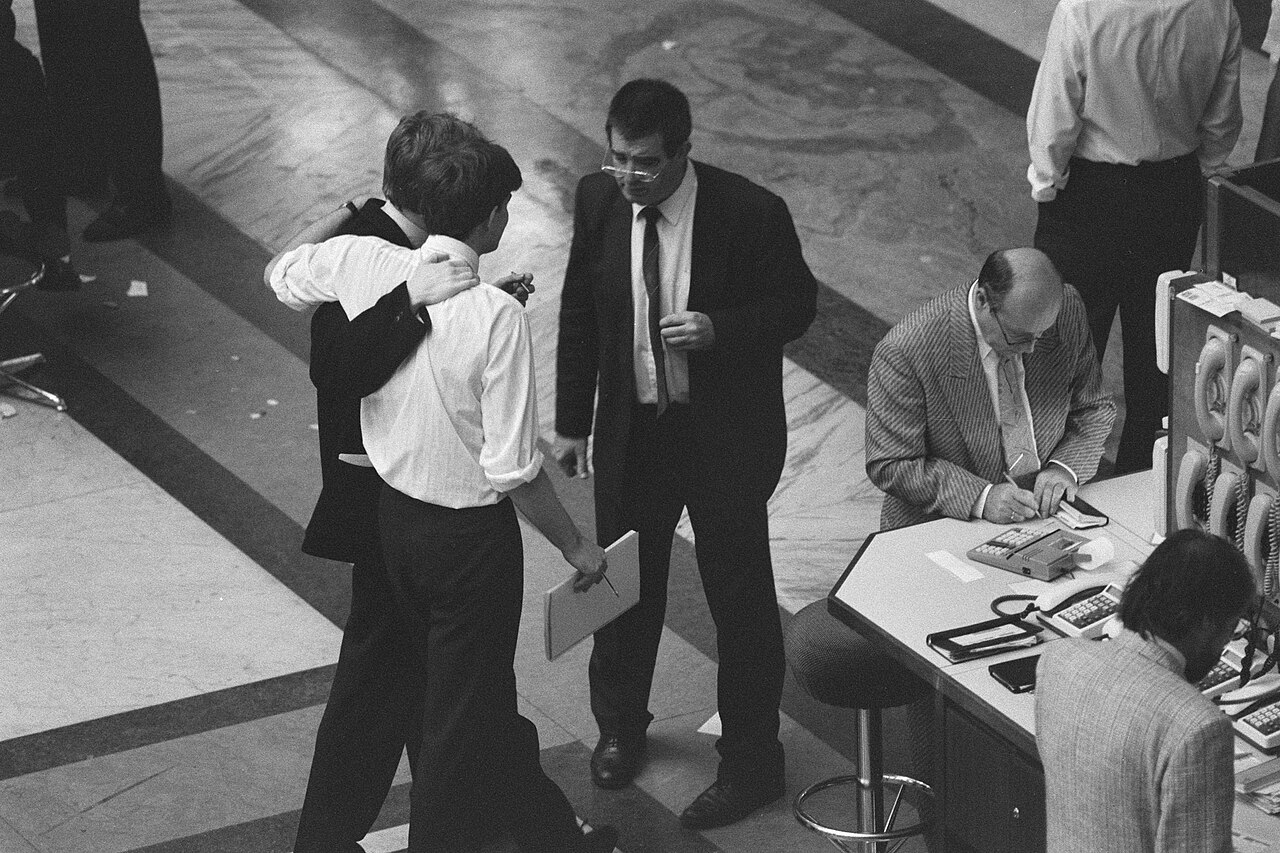 Vue plongeante sur un parquet de bourse. Un homme en bras de chemise passe le bras autour des épaules d'un collègue ; d'autres opérateurs consultent des papiers près d'un poste téléphonique.