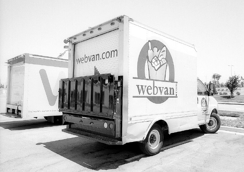Deux camions de livraison portant le logo « webvan.com » stationnés côte à côte sur un terrain vide, hayons ouverts.
