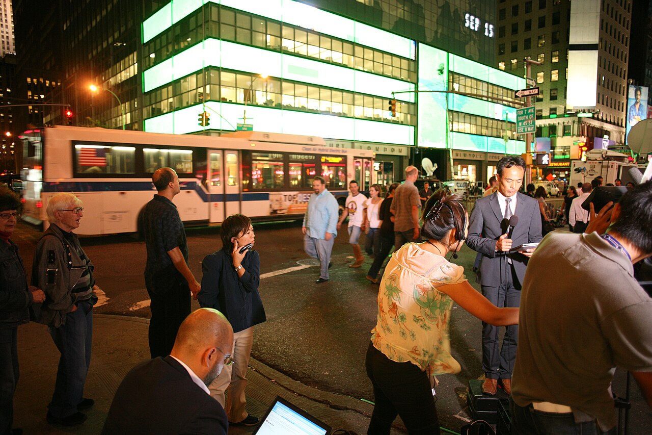 Une tour de bureaux vitrée éclairée la nuit ; un bandeau lumineux sur sa façade affiche « SEP 15 ». Sur le trottoir, des journalistes et des caméras sont installés.
