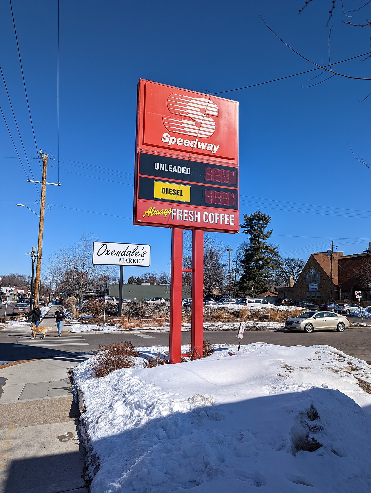 Un panneau de station-service au bord d'une route enneigée affiche en chiffres lumineux « unleaded 3,99 » et « diesel 4,99 ».