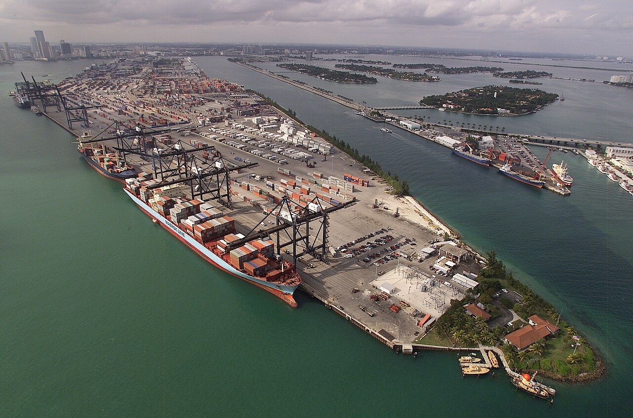 Vue aérienne d'un port : des milliers de conteneurs alignés en blocs le long des quais, des portiques de déchargement et un navire à quai.